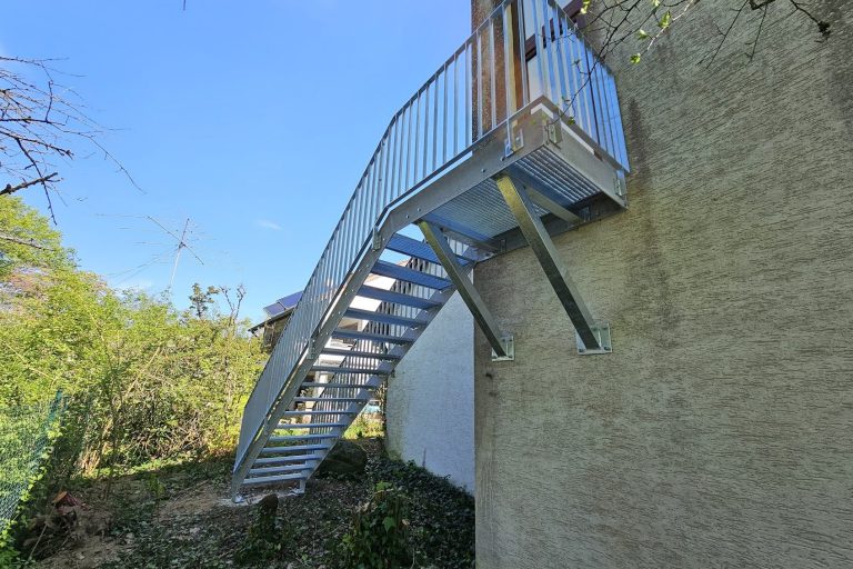Stahltreppe mit Podest und Geländer Eiserne Treppe, die an einer Wand nach draußen führt, umgeben von Bäumen und Sträuchern.